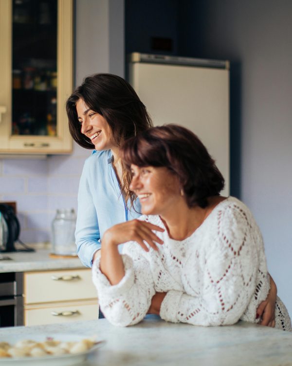 Two women smiling together in a kitchen, one leaning on the other's shoulder
