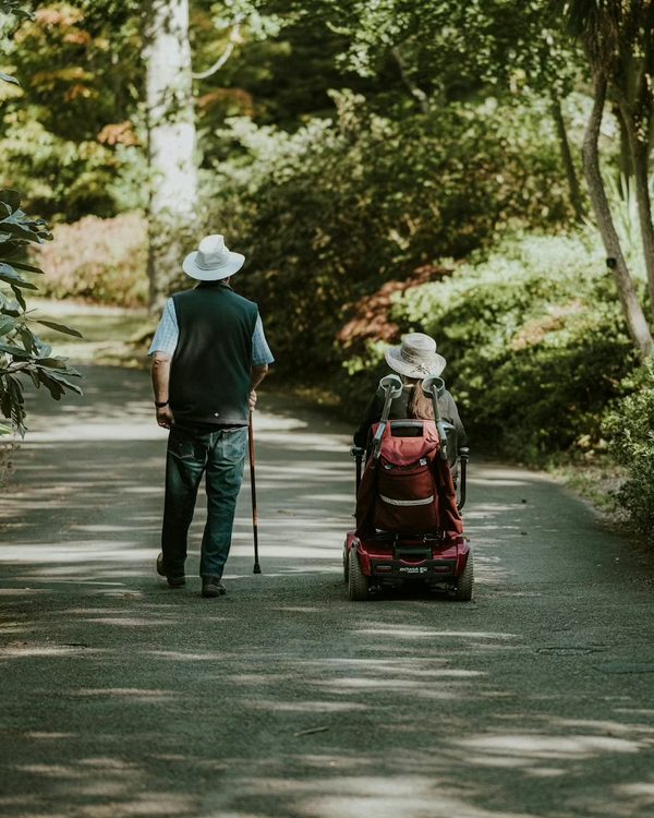 Two people walking a wooded park path together, one using a mobility scooter