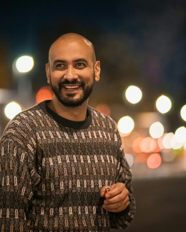 A man smiling on a city street at dusk with soft lights behind him